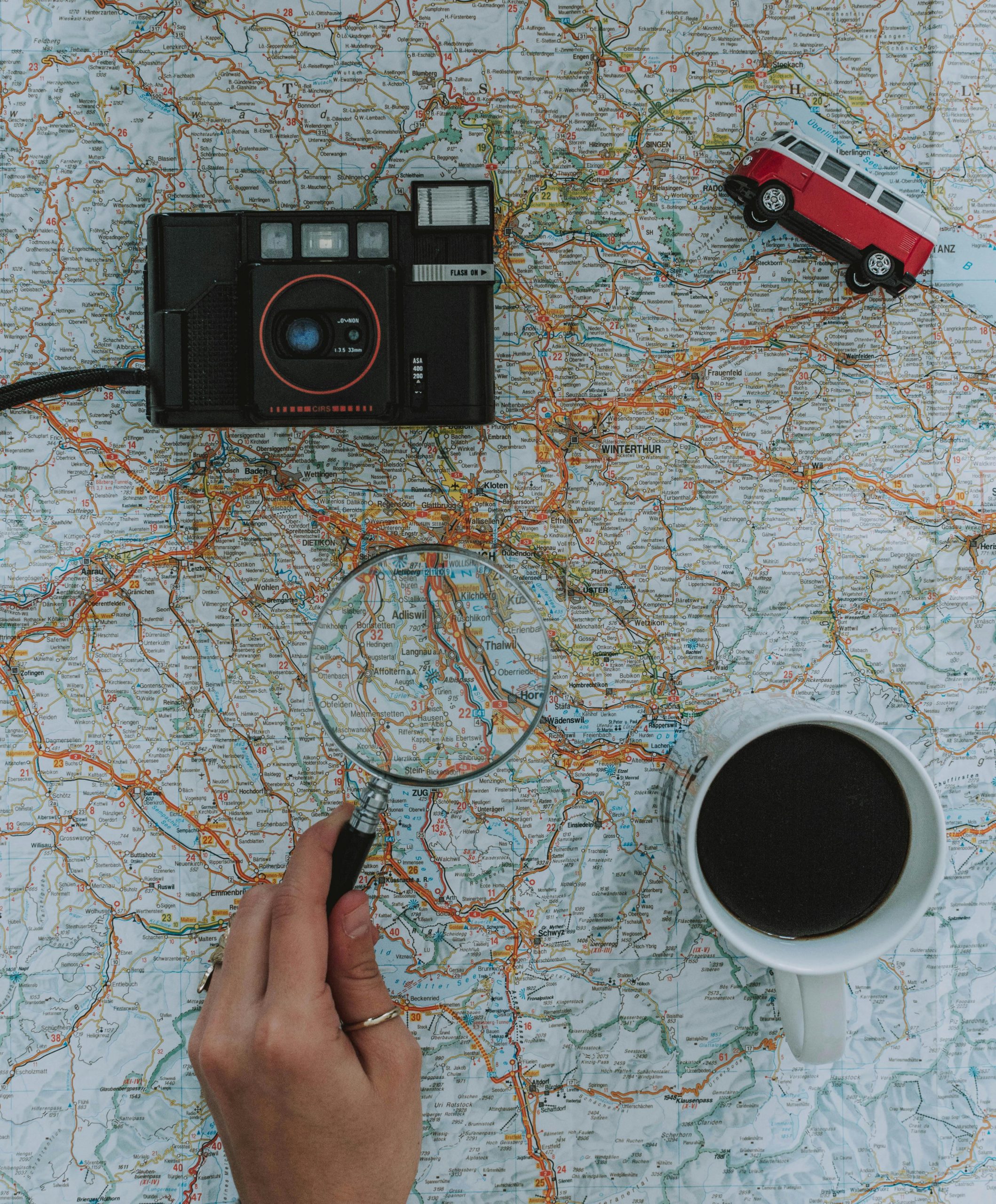 Flat lay of travel essentials: map, camera, toy van, magnifying glass, and coffee.