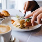Close-up of a person slicing a pastry with a cup of coffee on a white plate.