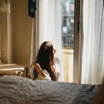 A woman sitting by a window in a sunlit bedroom, lost in thought and gazing outside.