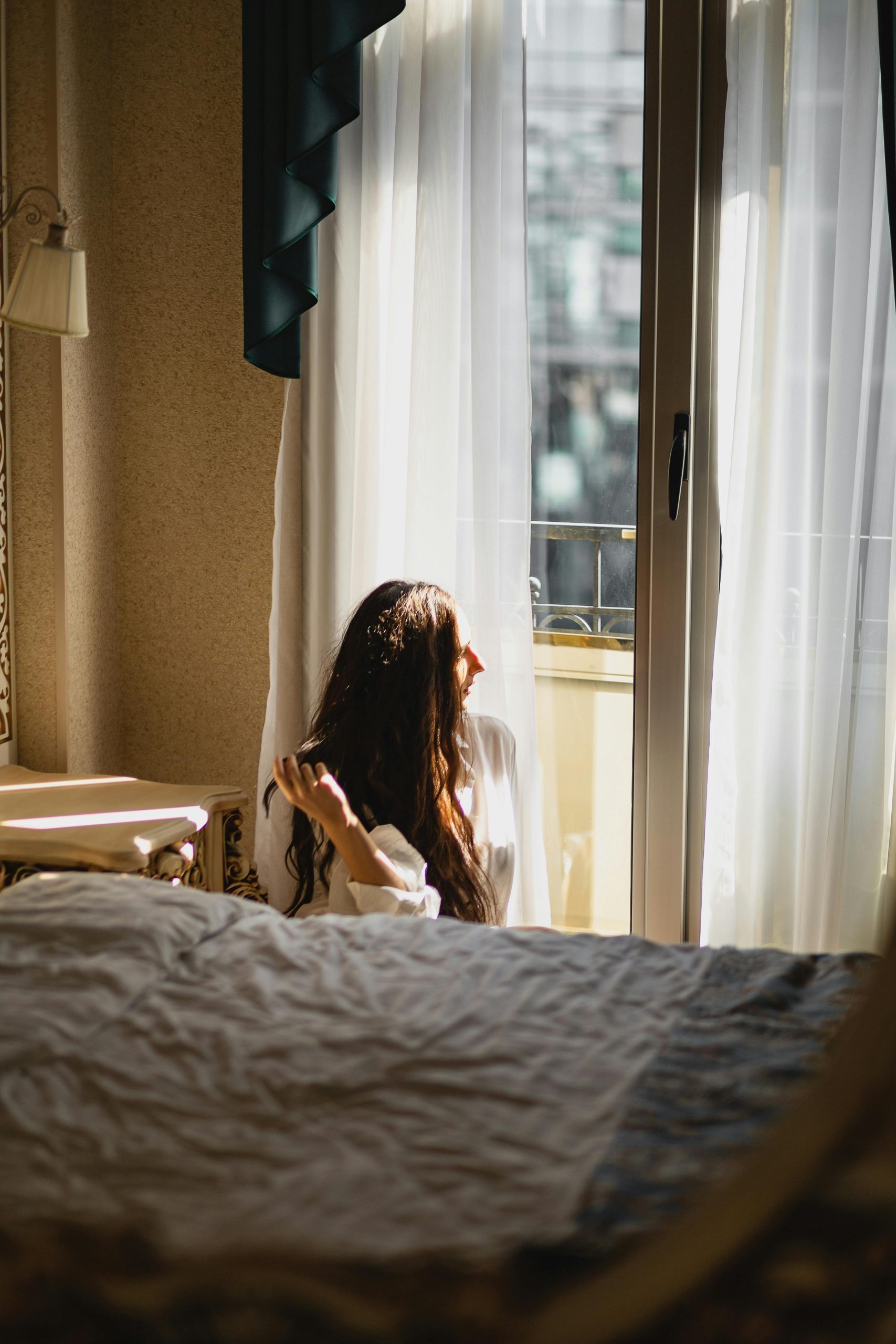 A woman sitting by a window in a sunlit bedroom, lost in thought and gazing outside.